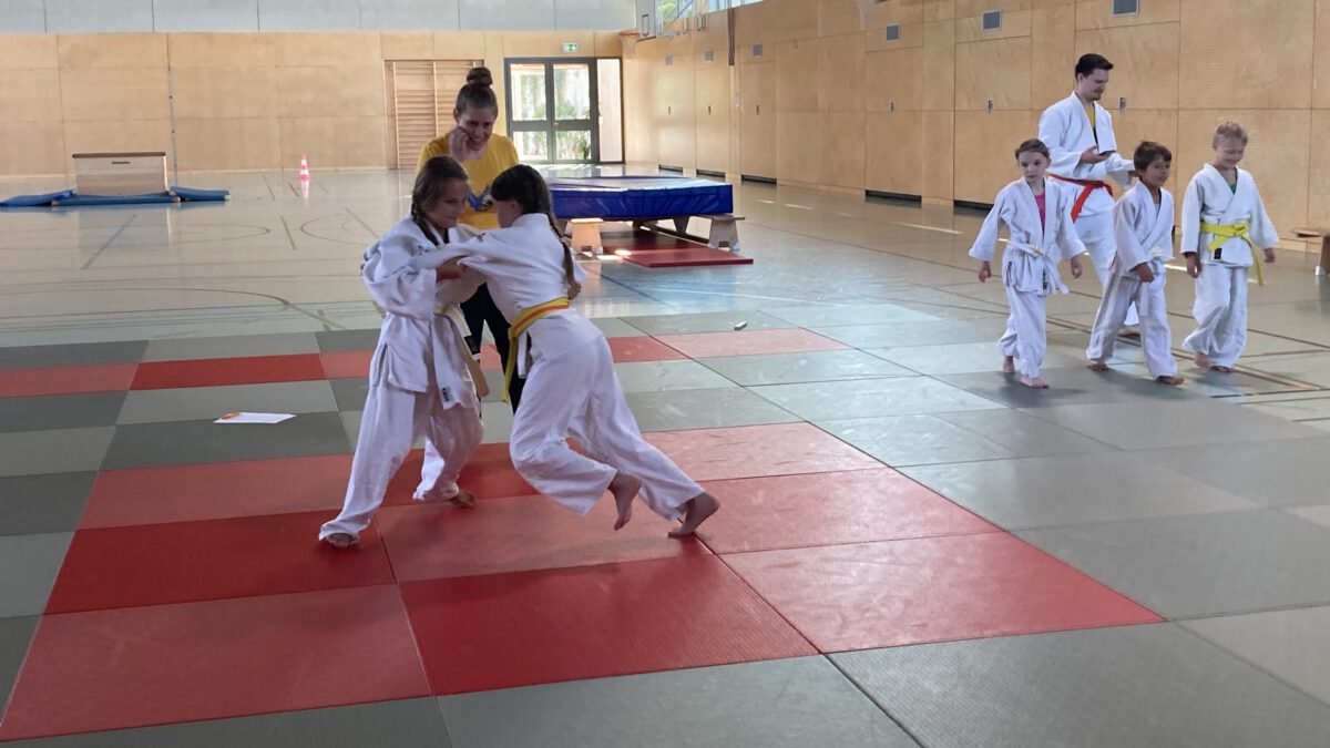Zwei Kinder, die eine Judo-Kampf machen. Dahinter die Trainerin, die zuschaut. Rechts im Hintergrund weitere Judokas.