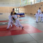 Zwei Kinder, die eine Judo-Kampf machen. Dahinter die Trainerin, die zuschaut. Rechts im Hintergrund weitere Judokas.