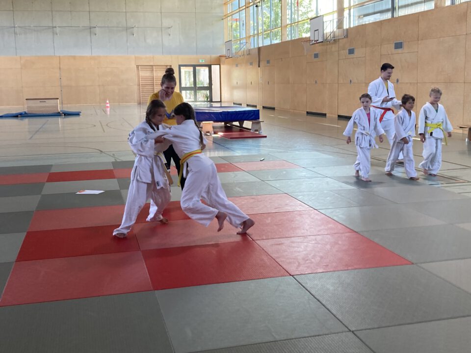 Zwei Kinder, die eine Judo-Kampf machen. Dahinter die Trainerin, die zuschaut. Rechts im Hintergrund weitere Judokas.